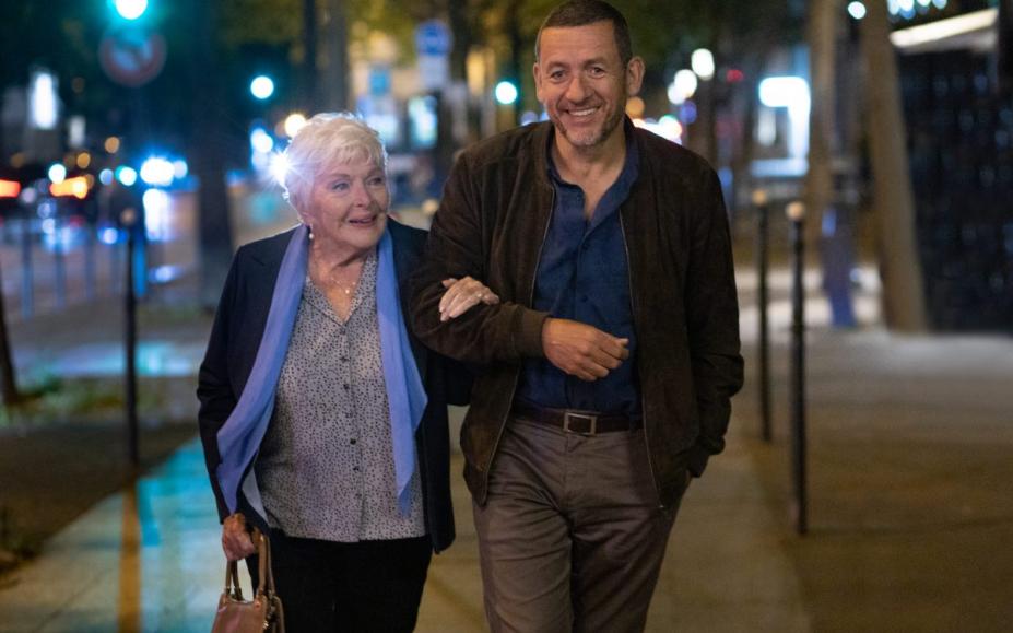 Mulher idosa e homem mais novo caminham de braços dados por uma rua à noite. #paratodosverem 