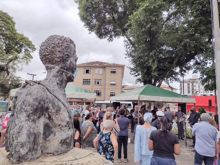 Busto de Zumbi dos Palmares, de máscara, com público em volta acompanhando cerimônia. #pracegover