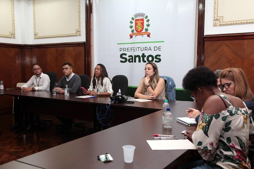 reunião com sete pessoas sentadas em torno de uma mesa. Ao fundo, um banner com o brasão da prefeitura. #paratodosverem