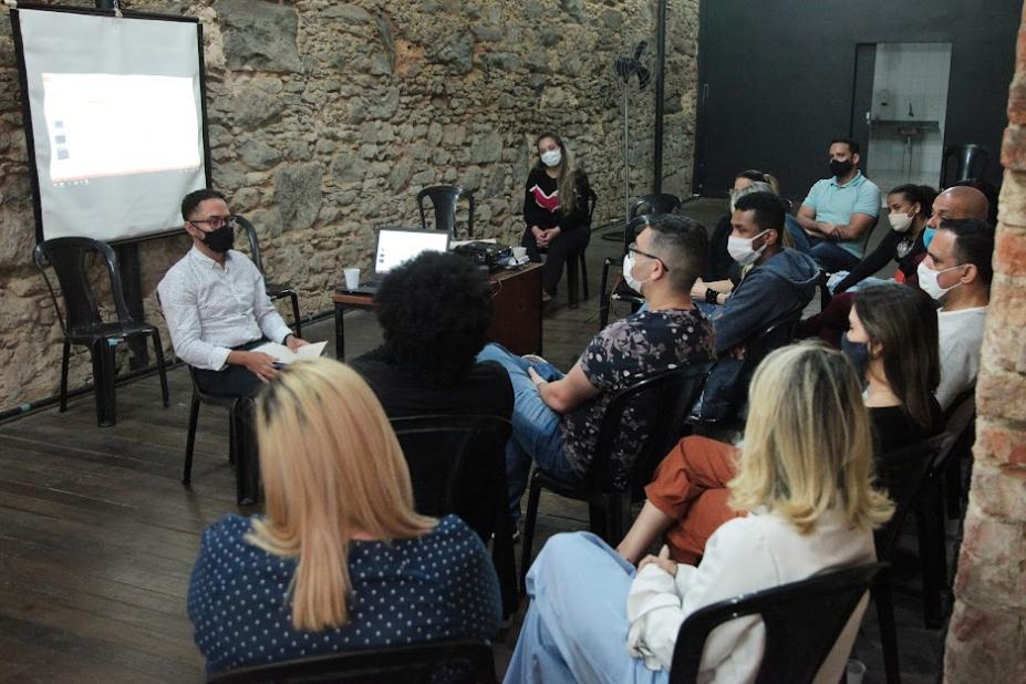 reunião em sala. Um homem está sentado falando a várias pessoas que estão sentadas diante dele. #paratodosverem