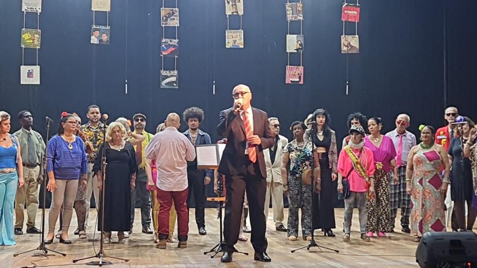 coral canta em palco. Integrantes estáo vestidos com roupas antigas. #paratodosverem 