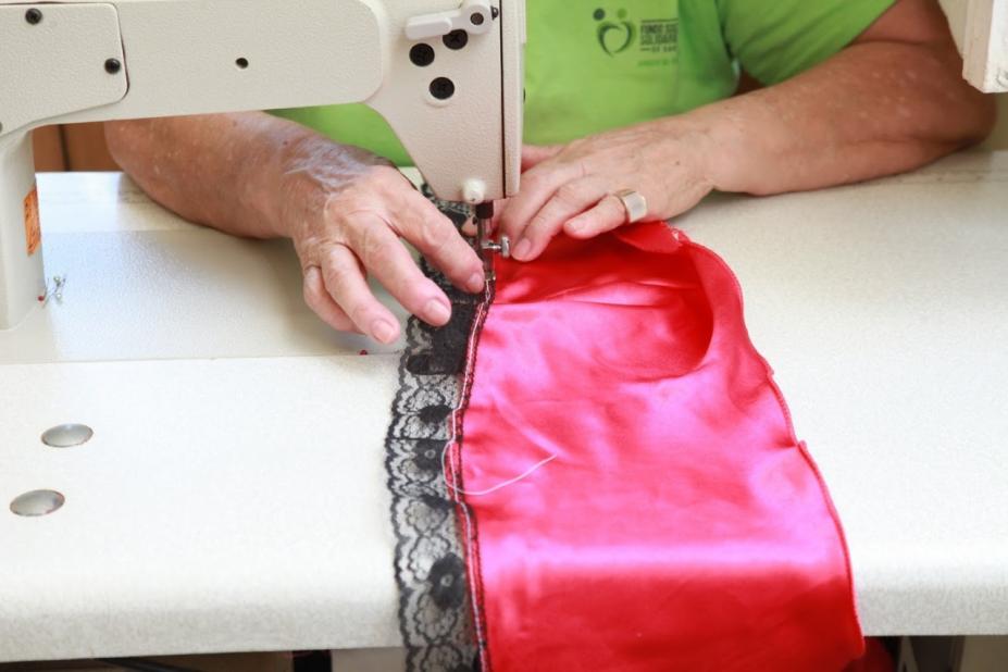 Imagem em close. Mãos de mulher seguram peça de roupa com renda em máquina de costura. #Paratodosverem