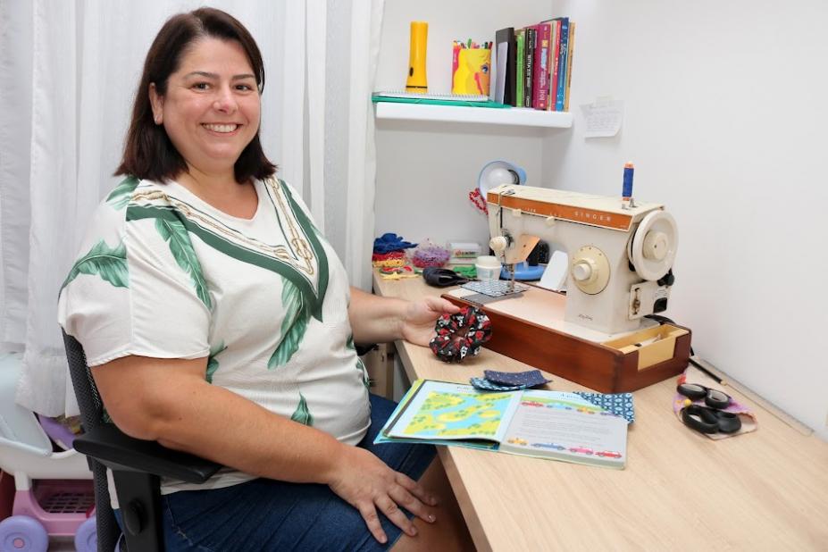 Alessandra está sentada diante de bancada onde está máquina de costura. Ela sorri para a foto. #paratodosverem