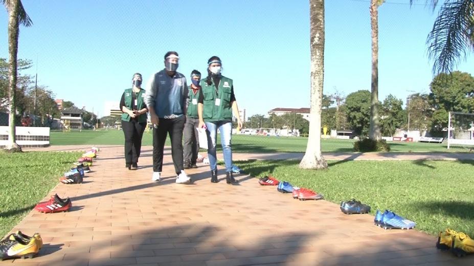 Agentes de força-tarefa estão em área aberta, um jardim. Nas laterias há chuteiras de futebol. #Paratodosverem