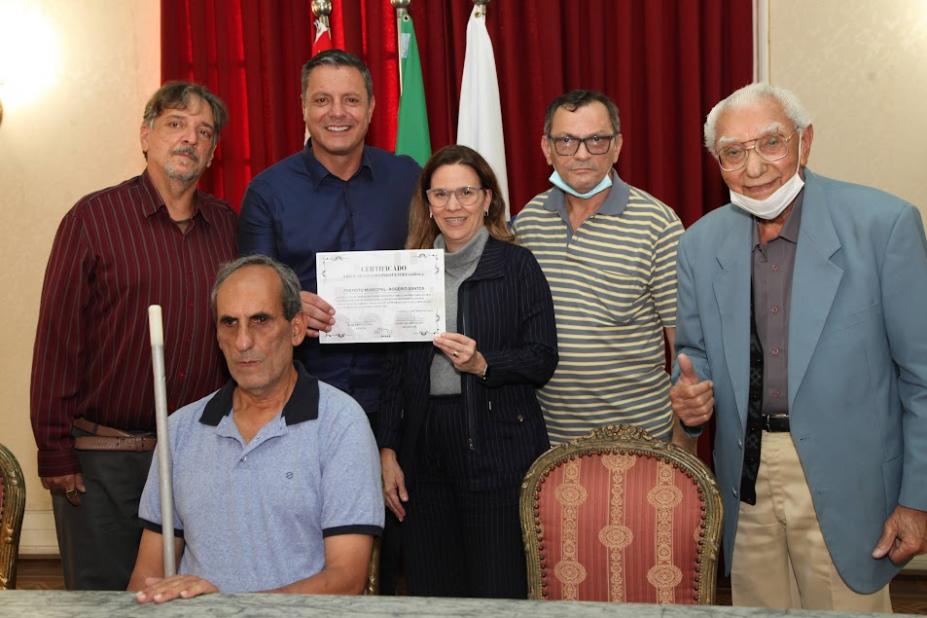 seis pessoas posam para foto. Cinco estão em pé. São representantes do setor cultural ladeando o prefeito e a vice-prefeito. Sentado, outro representante do setor cultura. Ele segura uma bengala branca. #paratodosverem