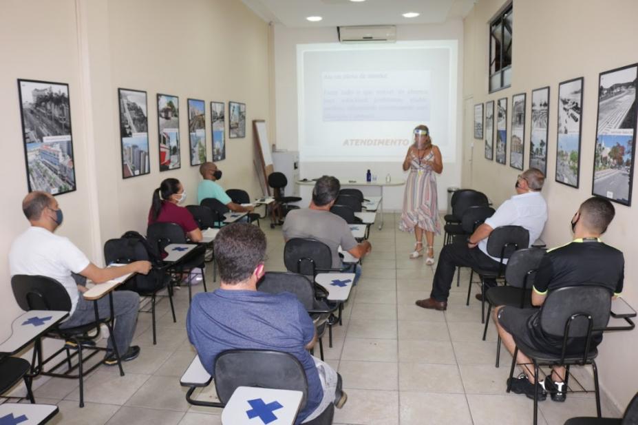 Pessoas sentadas em uma sala observam mulher falando a frente #paratodosverem