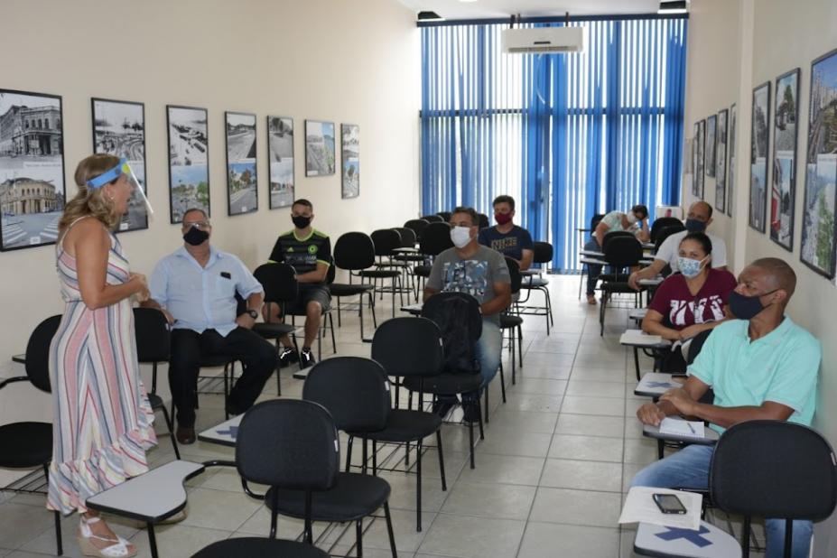 Sala de aula com professora em pé e alunos, adultos, sentados. #paratodosverem