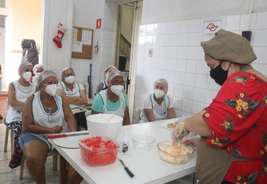 mulher prepara massa em recipiente de vidro sobre uma mesa. Outras mulheres sentadas assistem. #paratodosverem