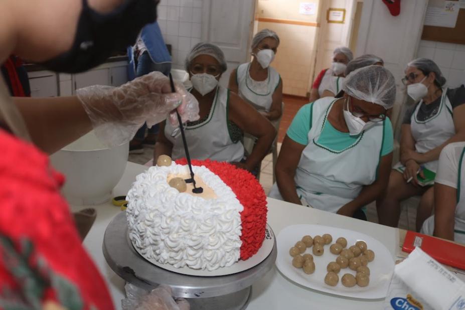 close em mãos que formam o rosto do papai noel sobre o chantilly do bolo. Alunas estão ao fundo observando e fazendo anotações. #paratodosverem