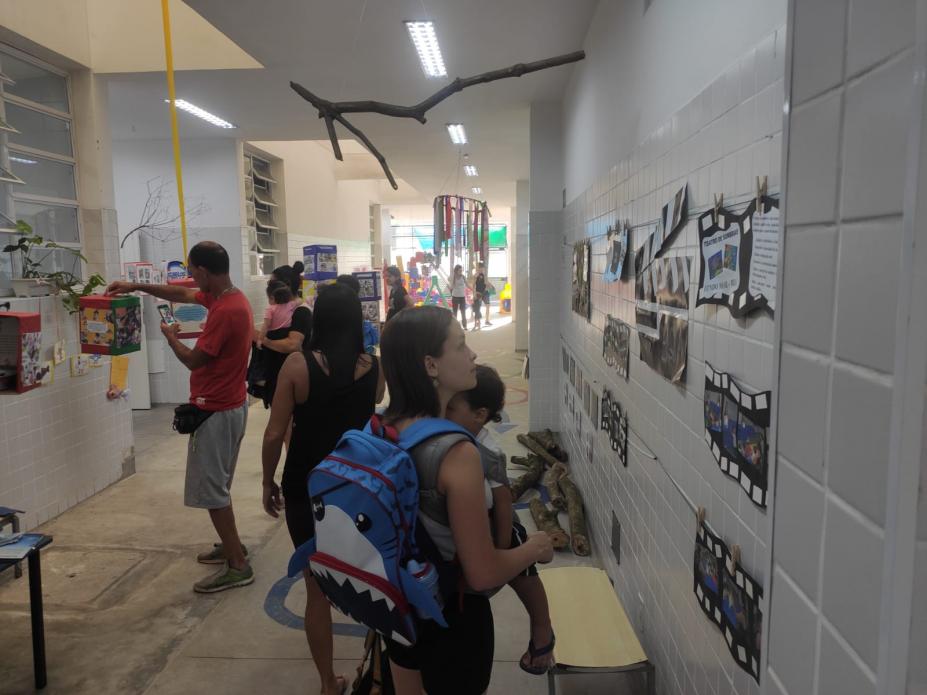 adultos olham fotos afixadas em parede de escola. #paratodosverem 