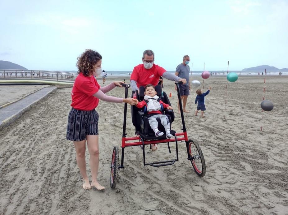 Criança com deficiência anda em bicicleta acessível, guiada por adultos, na faixa de areia. #paratodosverem