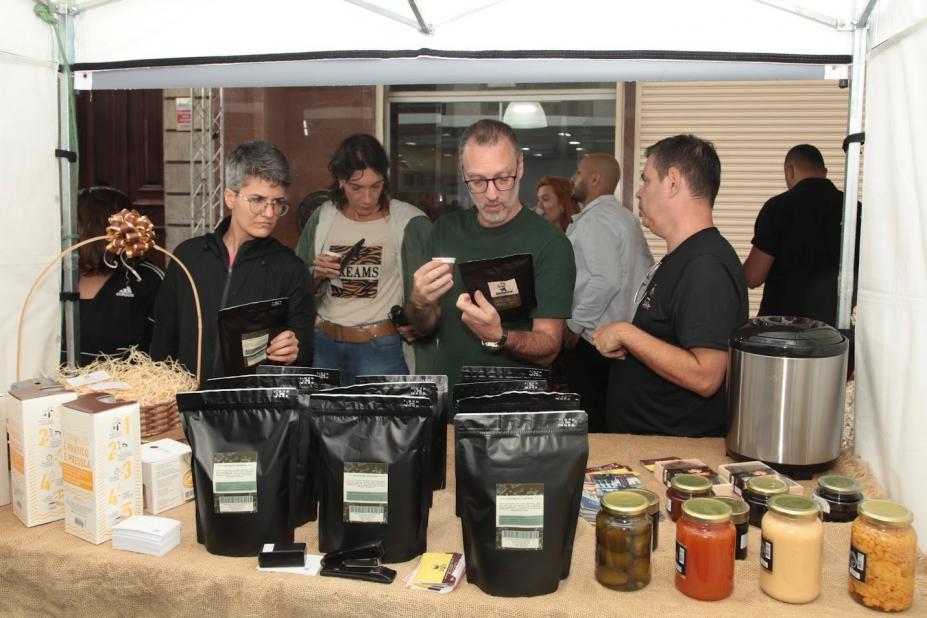 estande de café em primeiro plano, com pacotes do grão moído, e pessoas atrás, bebendo café. #paratodosverem 