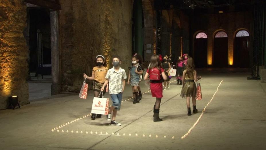 crianças desfilam em pista demarcada por luzes. Elas seguram sacolas. #paratodosverem