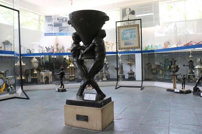 troféu no centro de uma sala com vários troféus espalhados. Peça tem dois homens segurando um troféu ao mesmo tempo. #paratodosverem