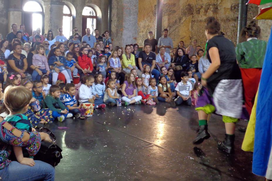 Dezenas de crianças acompanhadas de adultos assistem a apresentação de duas mulheres com roupas coloridas dentro da Casa da Frontaria Azulejada