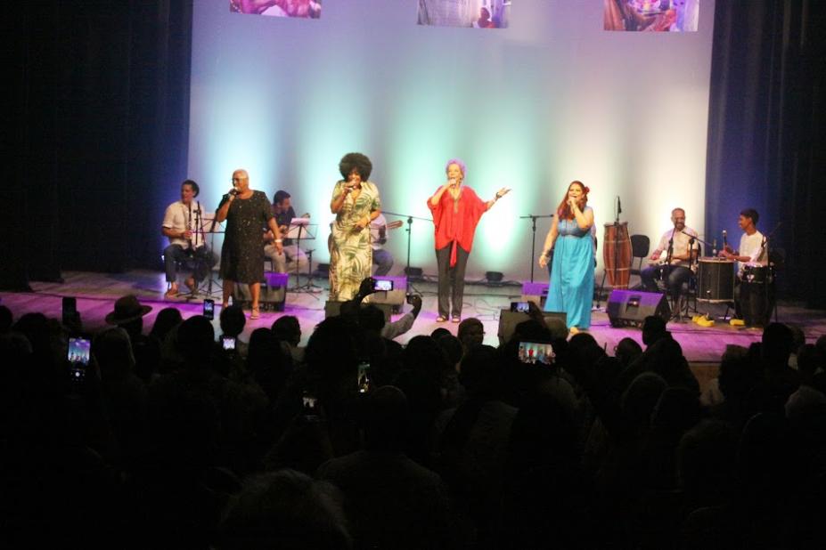 plateia lotada assiste mulheres cantando no palco. #paratodosverem