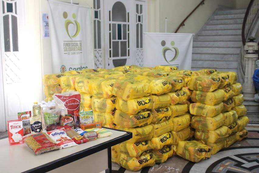 pacotes de alimentos sobre uma mesa e vários pacotes de cestas básicas apoiadas no chão e e empilhadas. A pilha deve ter pouco menos de 1,5 metro. #paratodosverem