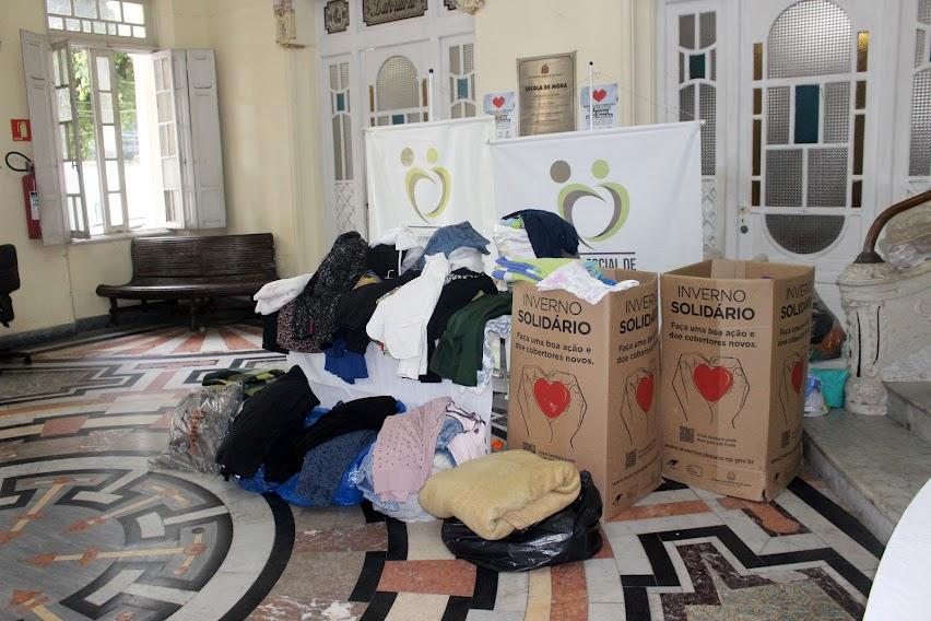 várias peças de roupas estão em caixas,  sobre uma mesa e no chão. Atrás, dois banners do fundo social de solidariedade