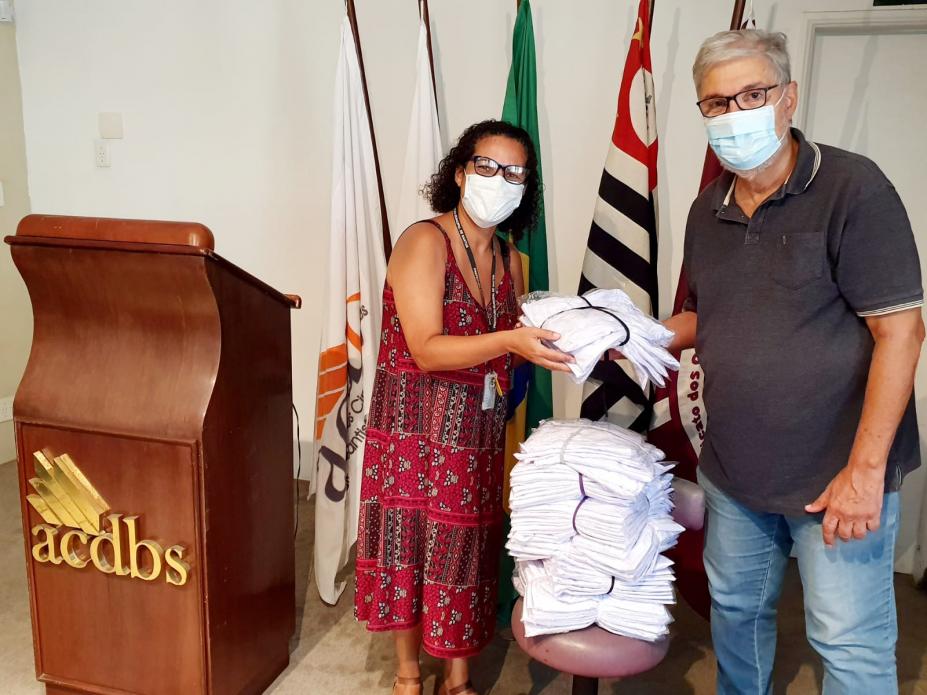 mulher e homem estão em pé, lado a lado, ambos segurando camisetas. Há uma pilha de camisetas sobre uma cadeira. Ao lado deles há uma tribuna onde estão as letras acdbs. #paratodosverem