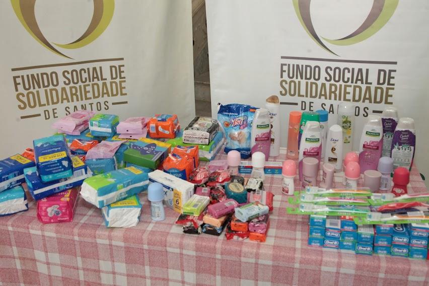 mesa com diversos itens de higiene. Atrás, dois banners do Fundo Social de Solidariedade