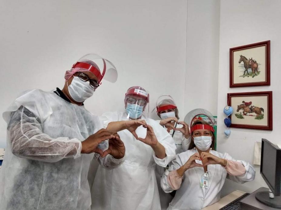 Quatro funcionários do setor de saúde estão usando avental, mascaras cirúrgicas e de acetato e gorro. Eles fazem gesto de coração com as mãos posando para a foto. #Paratodosverem