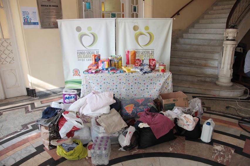 produtos de higiene e roupas estão sobre uma mesa e empacotados em torno dela. Atrás da mesa há um banner do fundo Social de solidariedade. #paratodosverem