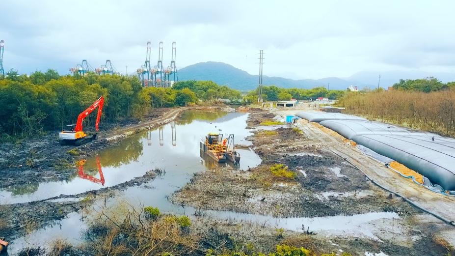 vista geral de canal máquinas para desassoreamento. Do lado esquerdo, vegetação. Ao fundo, guindastes do porto. #paratodosverem