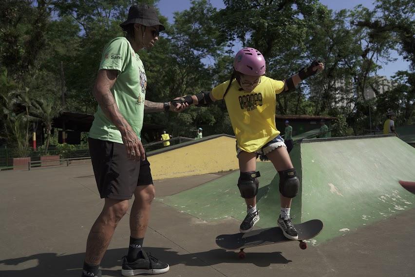 professor segura a mão de aluna que está sobre o skate. #paratodosverem