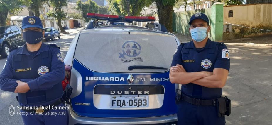 Dupla de guardas municipal, formada por uma mulher, à esq.) e um homem (à dir.), no centro, a viatura identificada com adesivo do programa Guardiã Maria da Penha. #paratodosverem