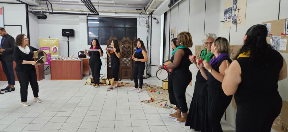 mulheres estão em semicírculo em pé tocando instrumentos de percussão. #paratodosverem