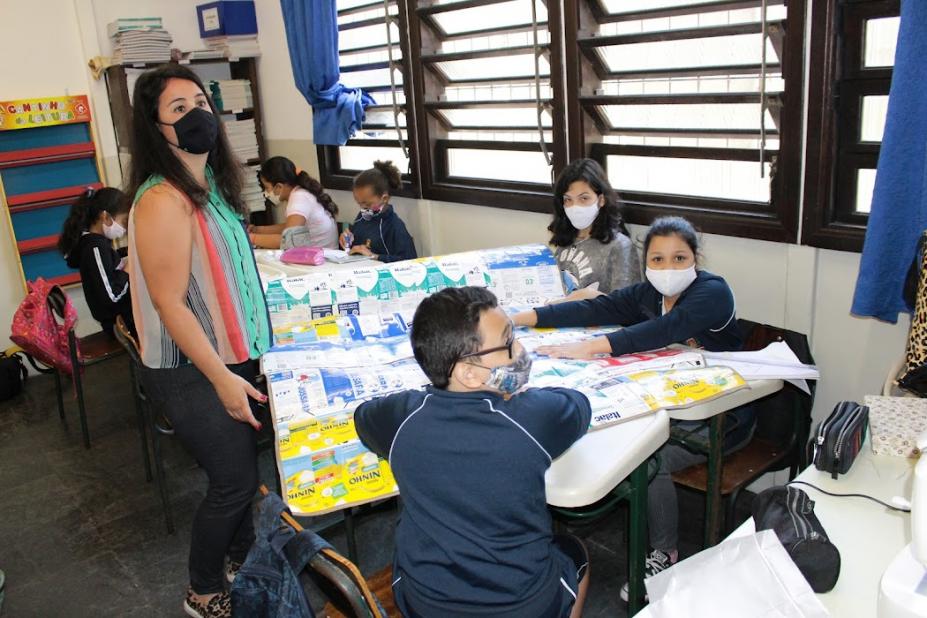 Professora e alunos na sala confeccionando cobertor com caixas de leite #paratodosverem