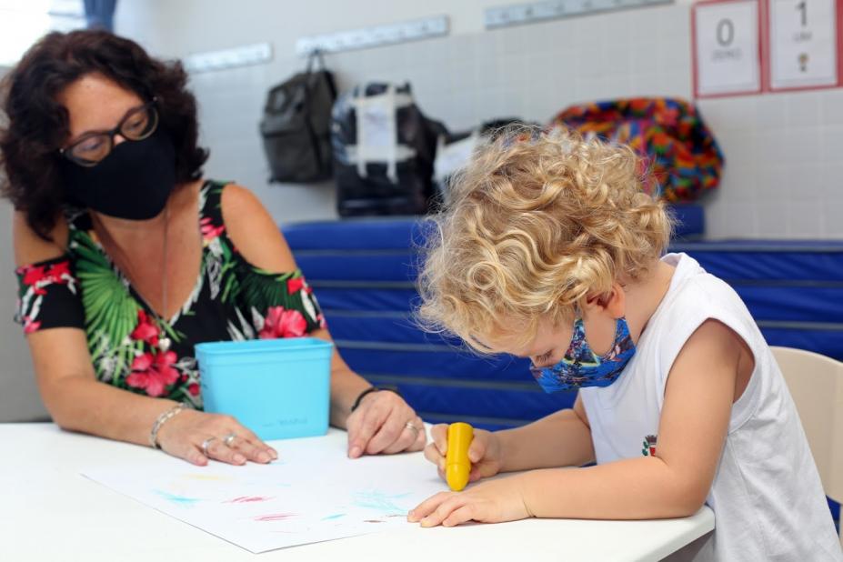 Educadora está ao lado de criança que desenha sobre uma mesa. #paratodosverem