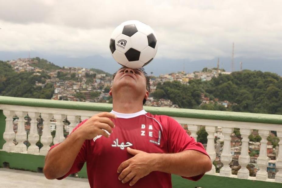 homem está equilibrando bola na cabeça.Ele está à frente de uma mureta no alto de um morro. Ao fundo, se vê as casas e a vegetação em outro morro. #paratodosverem