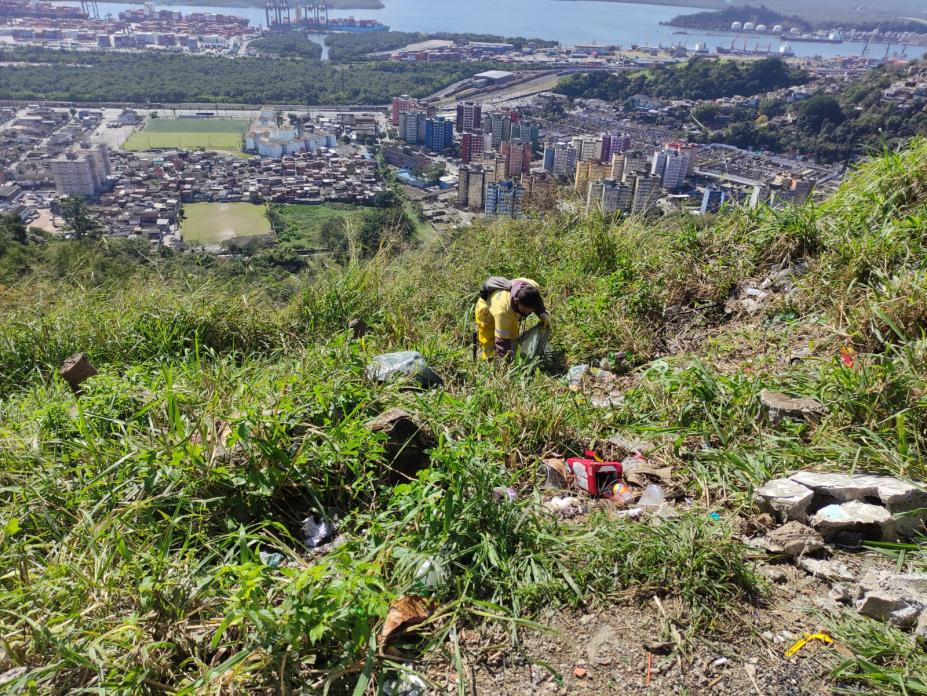 Homem retira lixo de área do morro com muita mata e cidade ao fundo. #paratodosverem