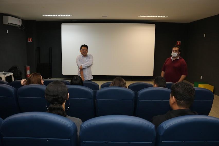 jovens estão sentados em poltrona de auditório atentes a dois homens que estão em pé falando. Ao fundo, um telão em branco. #paratodosverem