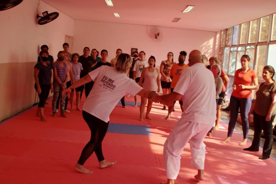 professor e professora fazem demonstração de movimento de defesa pessoa diante de várias mulheres. todos estão sobre um tatame. #paratodosverem 