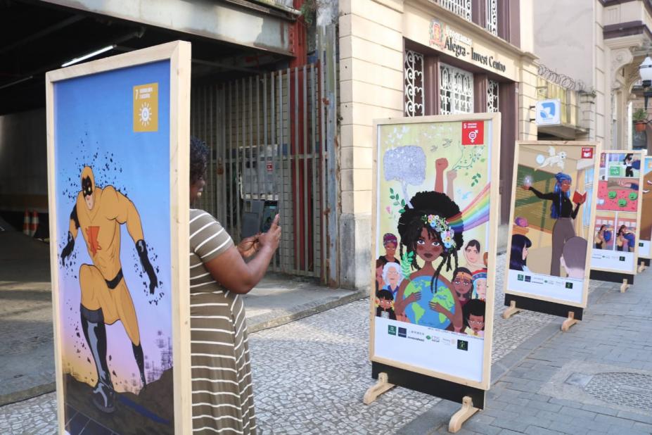 paineis expostos em calçadão. Atrás de um deles aparece a lateral do corpo de uma mulher que fotografa uma das obras. #paratodosverem 
