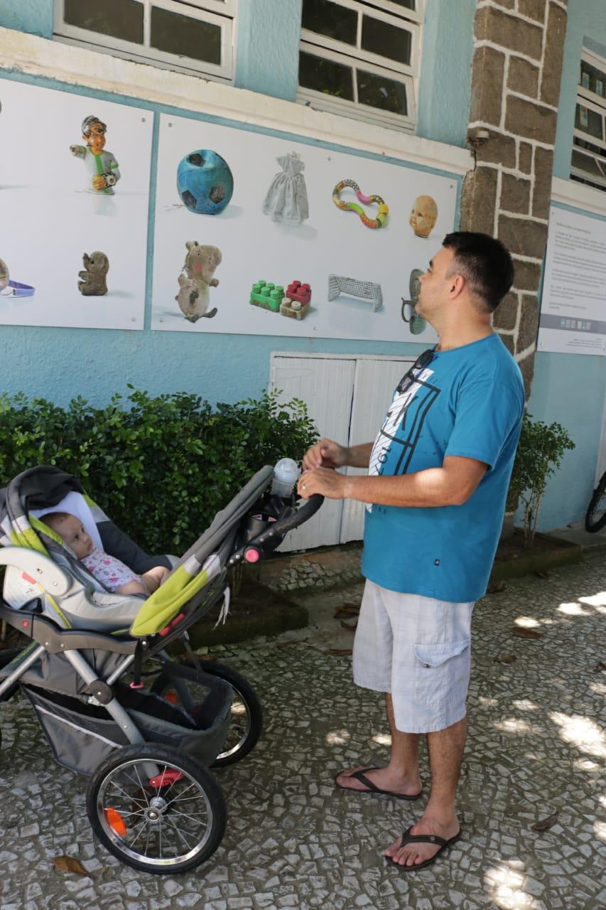 Homem com bebê ao lado em carrinho olha fotos da exposição. #pracegover