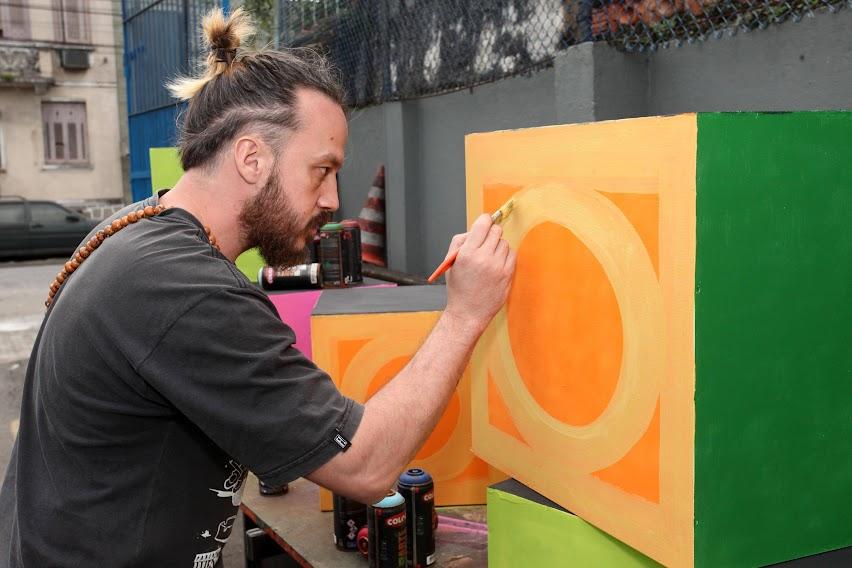 artista de rua está pintando um cubo com desenho da mureta tradicional de santos. #paratodosverem