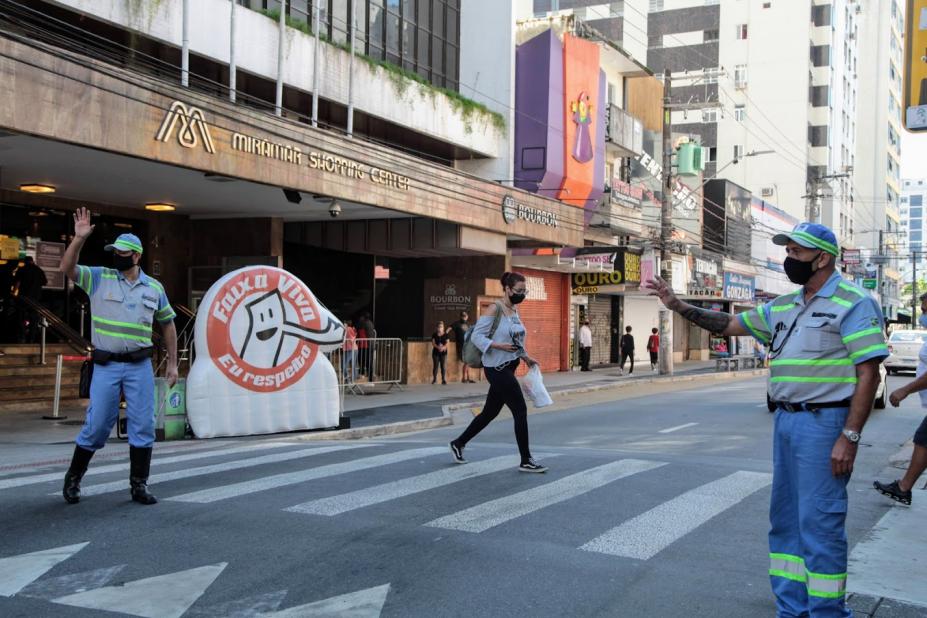 Agentes da CET fazem gesto de mão estendida sobre faixa de pedestre. Ao fundo, uma mulher atravessa a rua. Do lado esquerdo, o inflável da campanha Faixa Viva. #paratodosverem