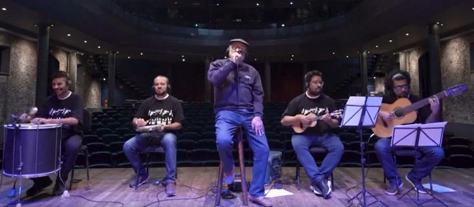 quatro músicos tocando instrumentos sentados e um ao centro cantando. #paratodosverem