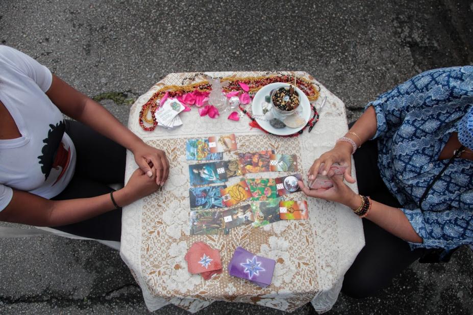 foto feita do alto mostra o tronco e as mãos de duas pessoas frente a frente em uma mesa. A da direita manuseia um baralho. #paratodosverem