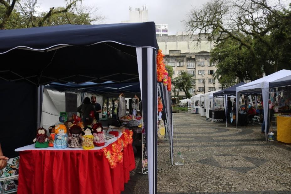 barraca de artesanato em primeiro plano e várias outras enfileiradas em praça. #paratodosverem