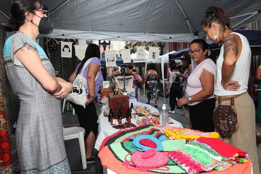 Mulheres observam produtos de crochê em estande. #pratodosverem