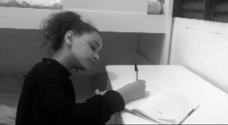 Menina está escrevendo em caderno ao lado de cama beliche. A foto em preto e branco dá um tom sombrio à cena. #Paratodosverem