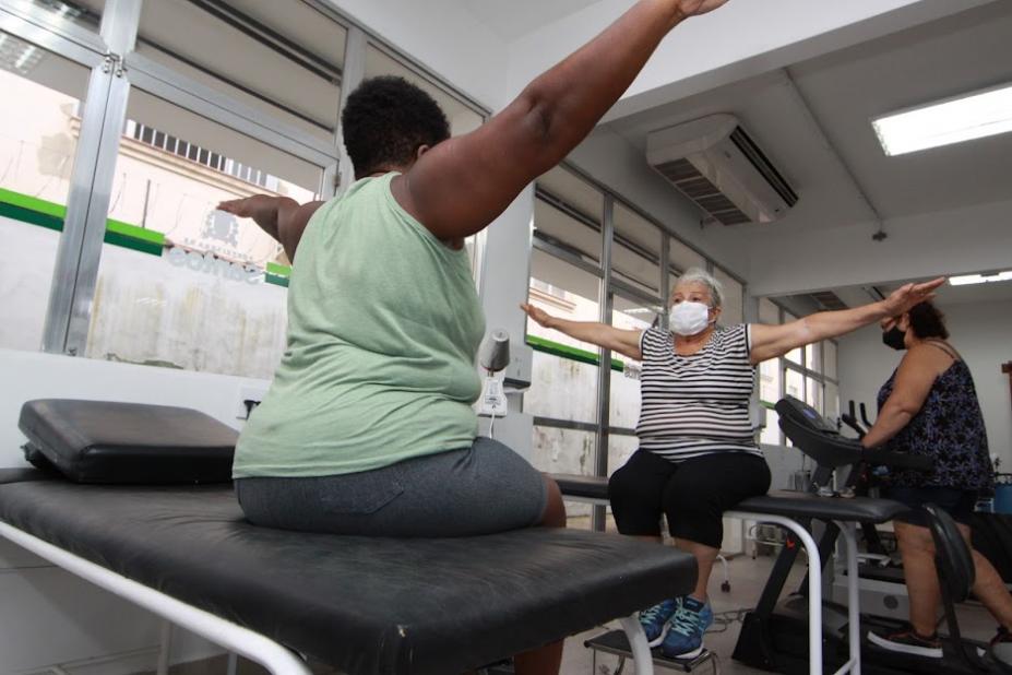 duas mulheres estão sentadas em macas fazendo movimentos com os braços. Eles estão estendidos nas laterais. #paratodosverem