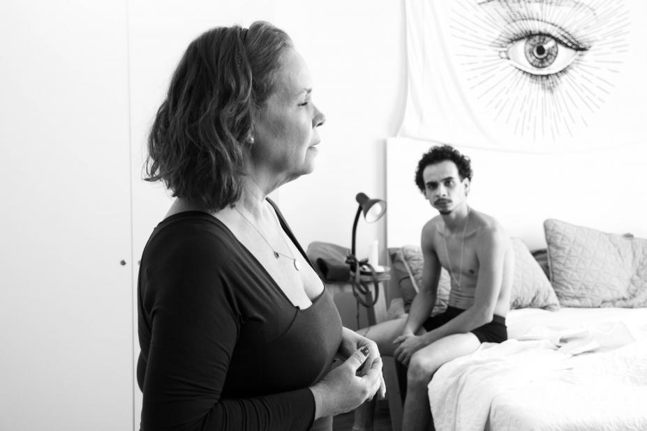 cewna de fim. Uma mulher, de lado, está em pé. Sem olhar diretamente, ela fala com um homem que está sentado em uma cama. #paratodosverem