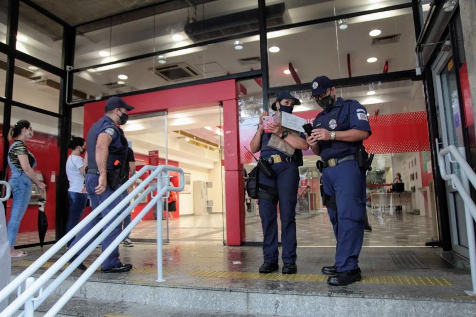 Agentes estão na porta de entrada de um banco. Eles têm bloco de anotações nas mãos. #Paratodosverem