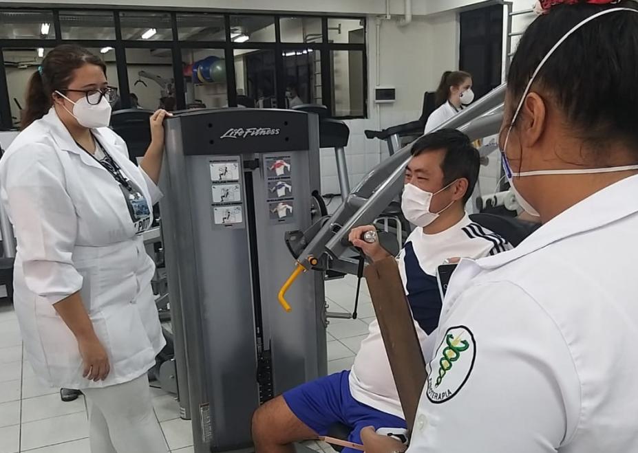 Estudantes orientam homem em exercício em aparelho de musculação. #pracegover