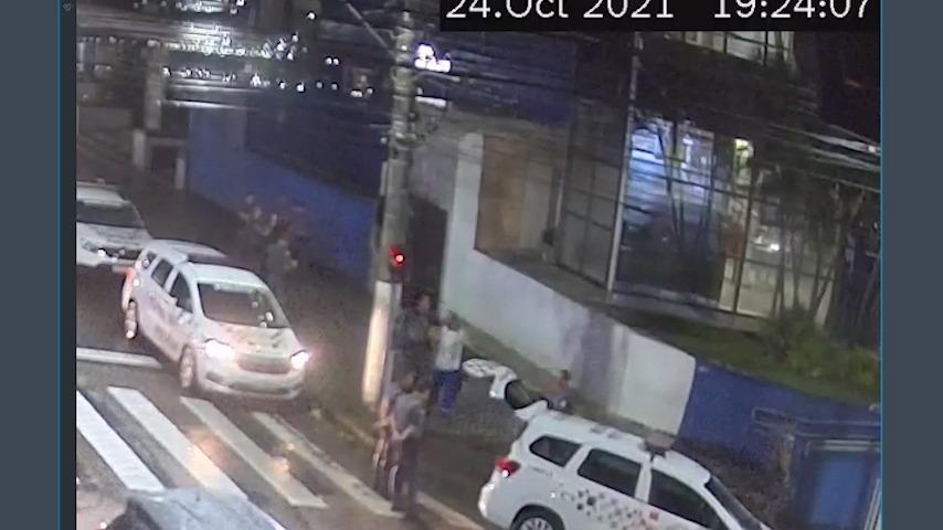 Duas viaturas da polícia militar estão paradas junto ao meio-fio. Um policial conduz um homem para dentro do carro. #paratodosverem